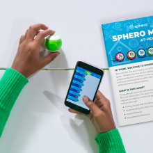Basılı öğrenme kılavuzunun yanında Sphero Mini oyuncak robot ve blok kodlu telefon tutan çocuk.